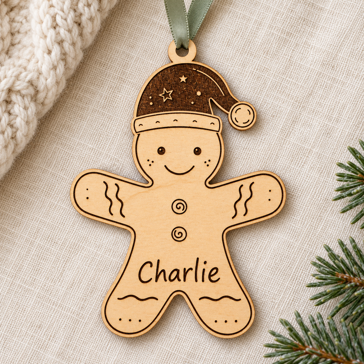 Named gingerbread Christmas ornament on ivory linen beside cream knit fabric and a fir branch