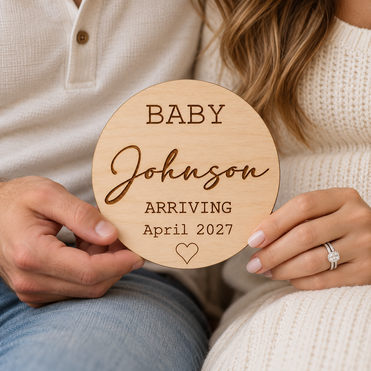 Parents holding a round Baby Johnson Arriving April 2027 announcement sign