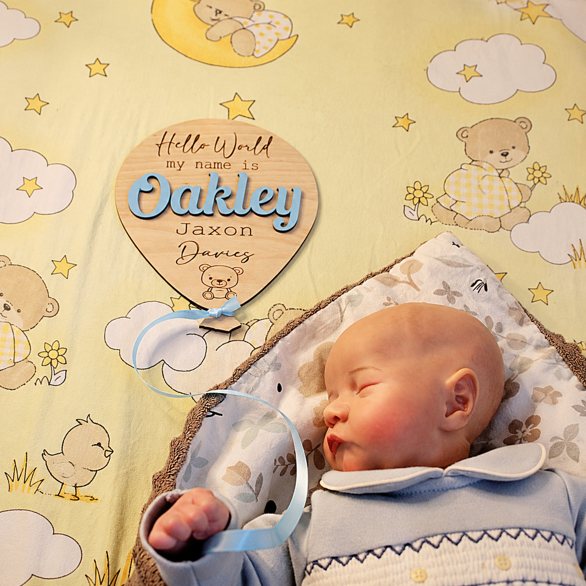 Oakley Jaxon Davies wooden birth announcement balloon with Blue satin ribbon beside a sleeping newborn on yellow nursery bedding.