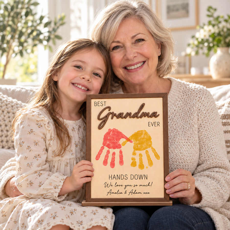 Grandmother and granddaughter holding a brown Best Grandma Ever wooden frame decorated with children's handprints.