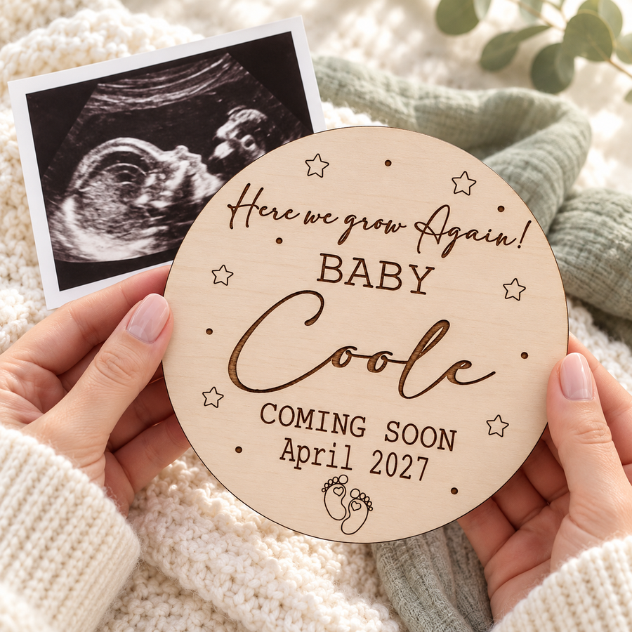 Hands holding a personalised Here We Grow Again wooden pregnancy announcement sign beside an ultrasound