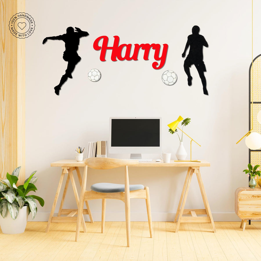 A football themed decoration with the name "Harry" in bright red letters, flanked by two black silhouettes of soccer players. There are also two soccer balls beneath the name. The sign is mounted on a white wall above a simple wooden desk with a computer. It has a handmade quality and would look great in a kid's bedroom or study area for a soccer fan.