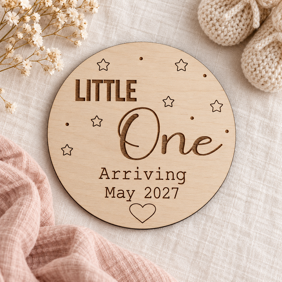 Little One Arriving May 2027 wooden plaque on ivory linen beside cream baby booties and blush muslin
