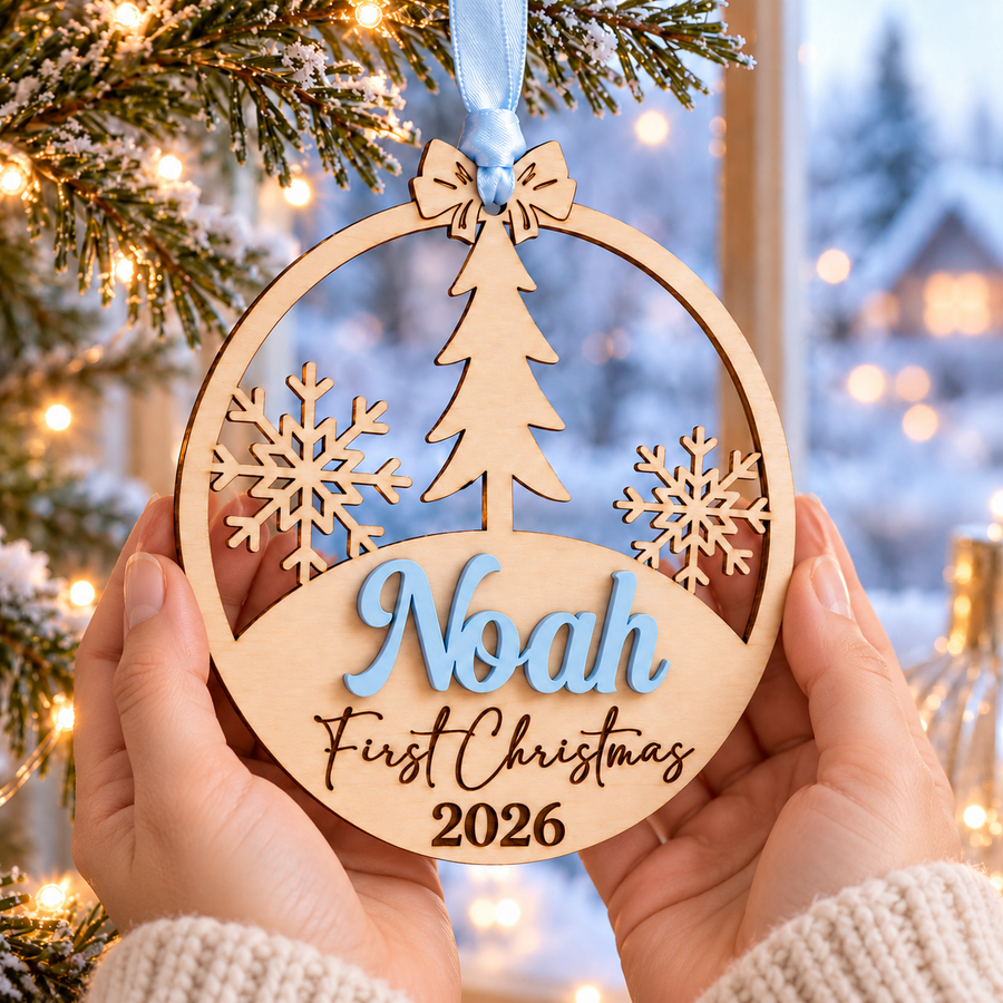 Personalised baby's first Christmas wooden bauble with Noah in raised pastel blue lettering and engraved 2026, held in adult hands against a festive winter background