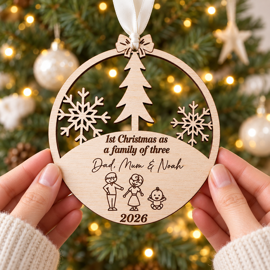 Personalised wooden first Christmas as a family of three bauble with Dad, Mum and baby Noah, held beside a Christmas tree