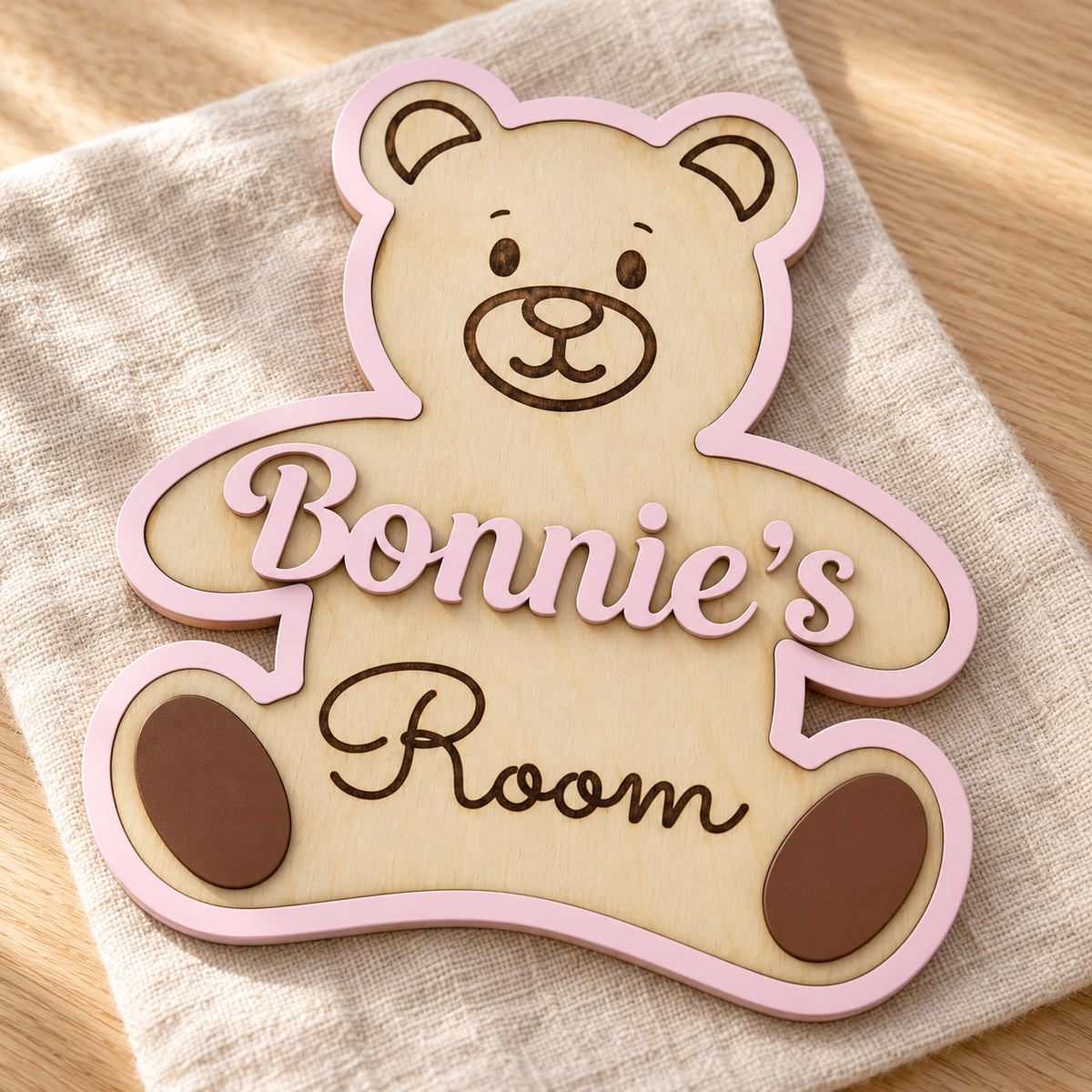 Close-up of raised pastel pink name lettering, natural birch wood layers and dark engraved details on a teddy bear room sign.
