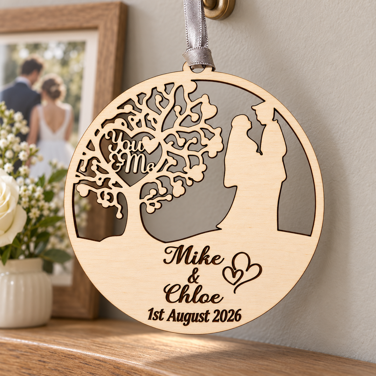 You and Me anniversary keepsake ornament hanging at home beside a wedding photograph and white flowers, with names and date engraved in birch.