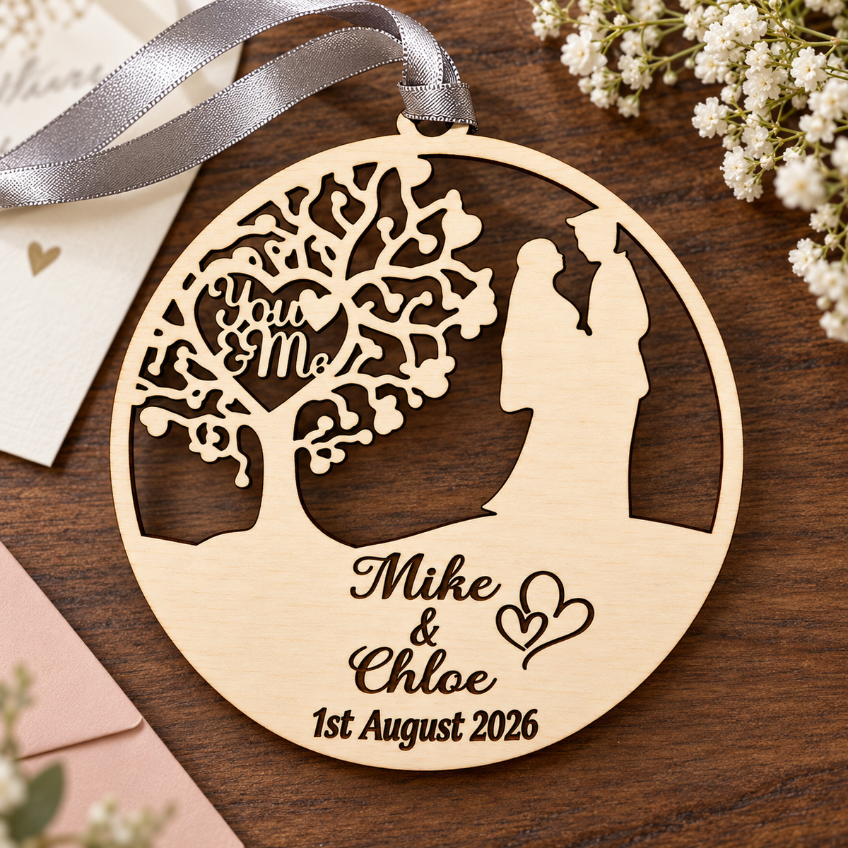 Natural wooden wedding gift ornament with engraved couple names and date, arranged on dark wood with a blush envelope and silver-grey ribbon.
