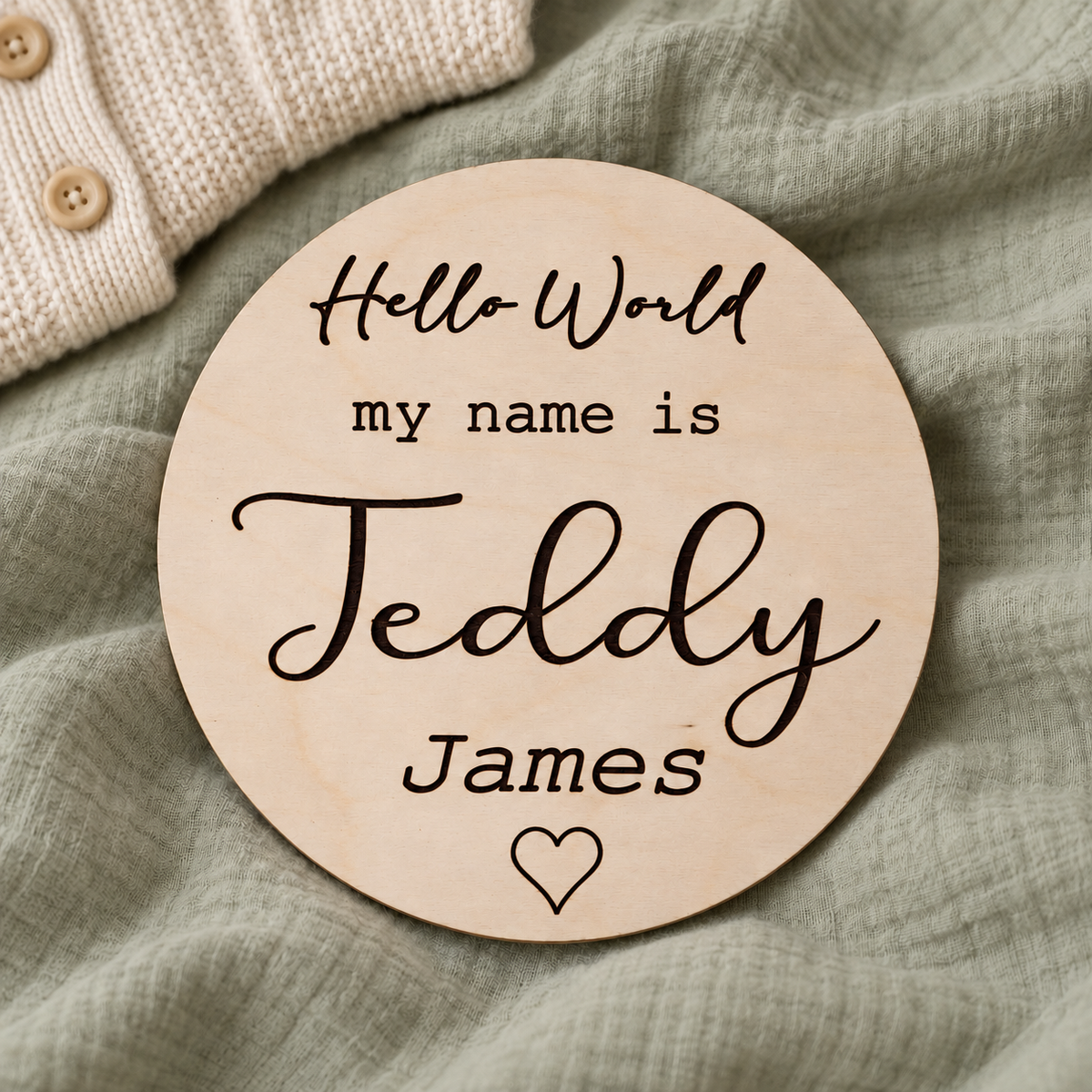 Round Hello World Teddy James birth announcement sign on pale sage muslin beside ivory knitwear