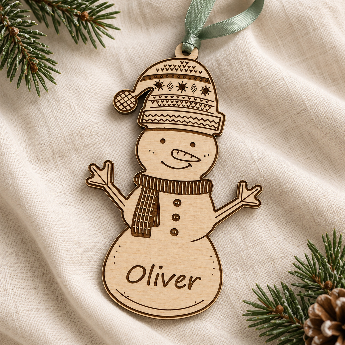 Birch plywood snowman Christmas decoration with an engraved name, patterned hat and sage ribbon on cream linen.