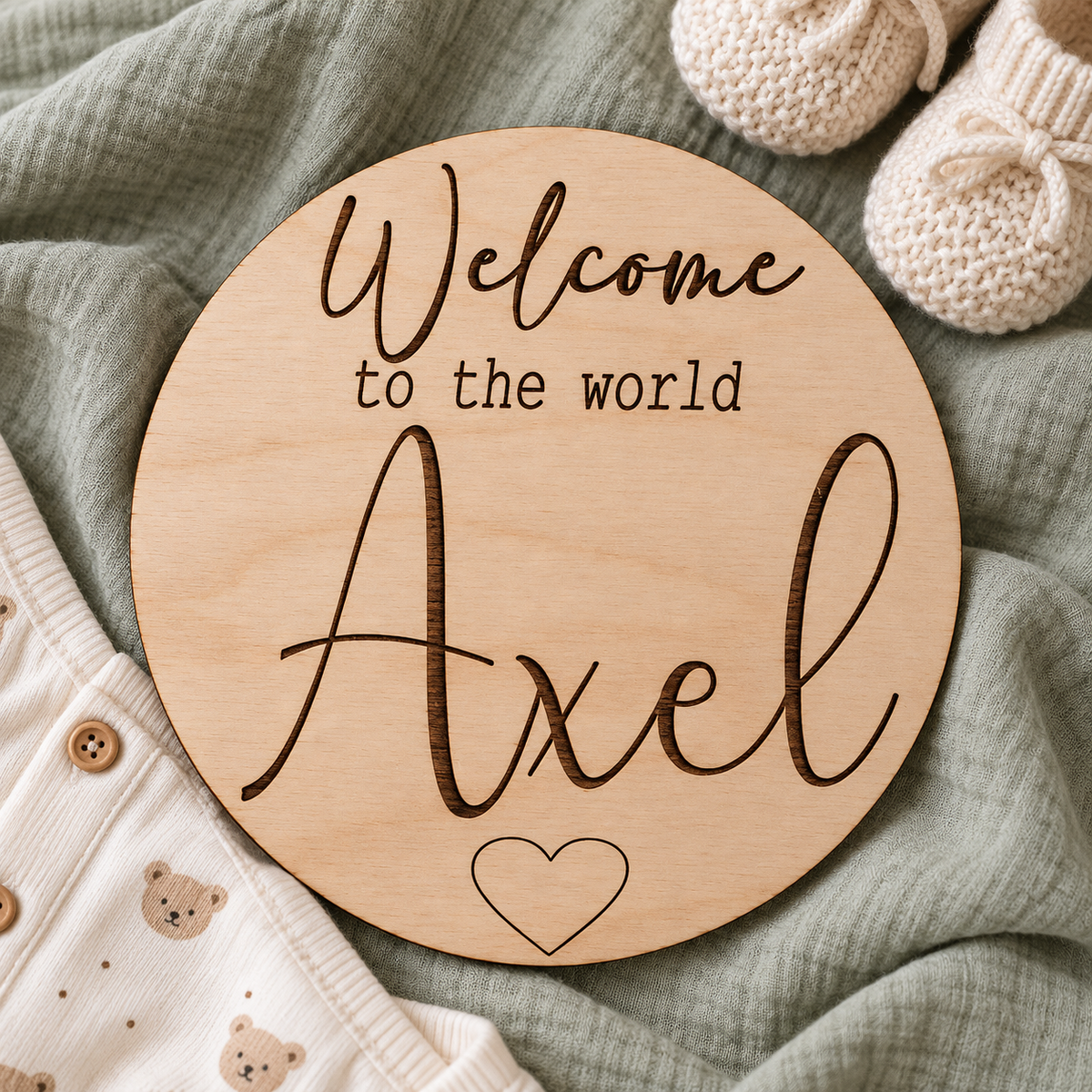 Round Welcome to the World Axel plaque on sage muslin beside ivory baby booties and a cotton outfit