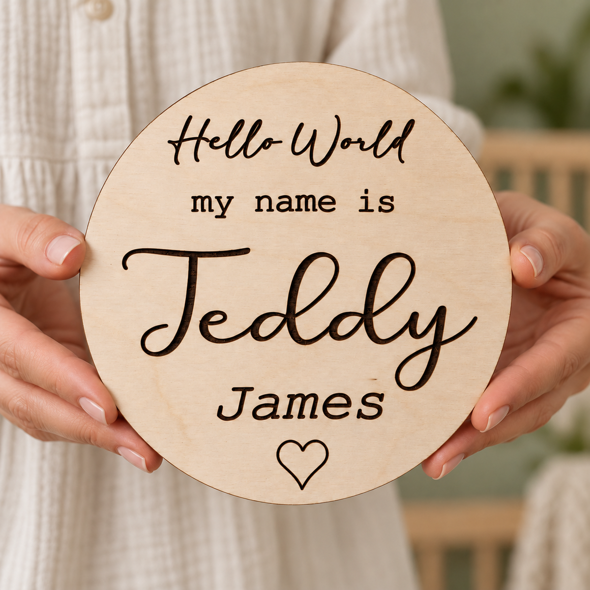 Hands holding a round birch birth announcement keepsake with dark Teddy James engraving