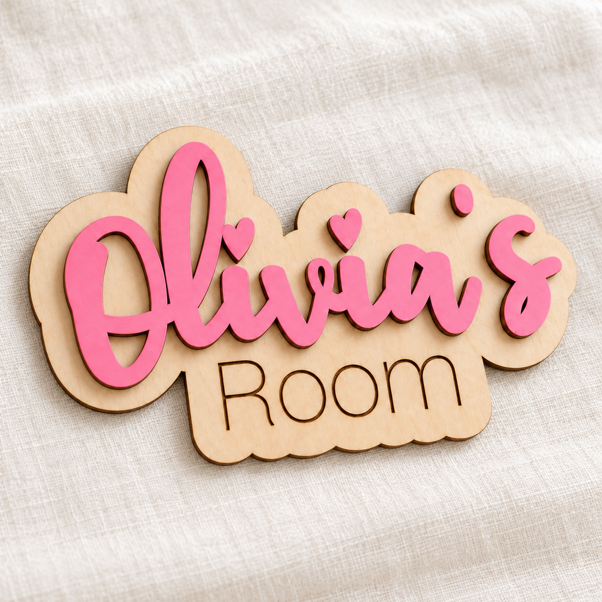 Close-up of pink raised Olivia's lettering and the engraved word Room on a natural birch plywood name sign