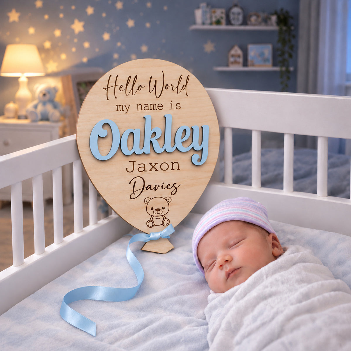 Oakley Jaxon Davies wooden baby announcement balloon with Pastel Blue raised name and Blue ribbon beside a newborn in a blue nursery.