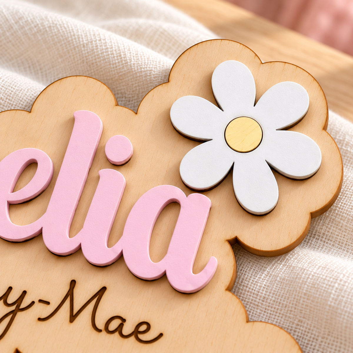Close-up of a white daisy with yellow centre, pastel pink raised wooden lettering and natural birch grain.