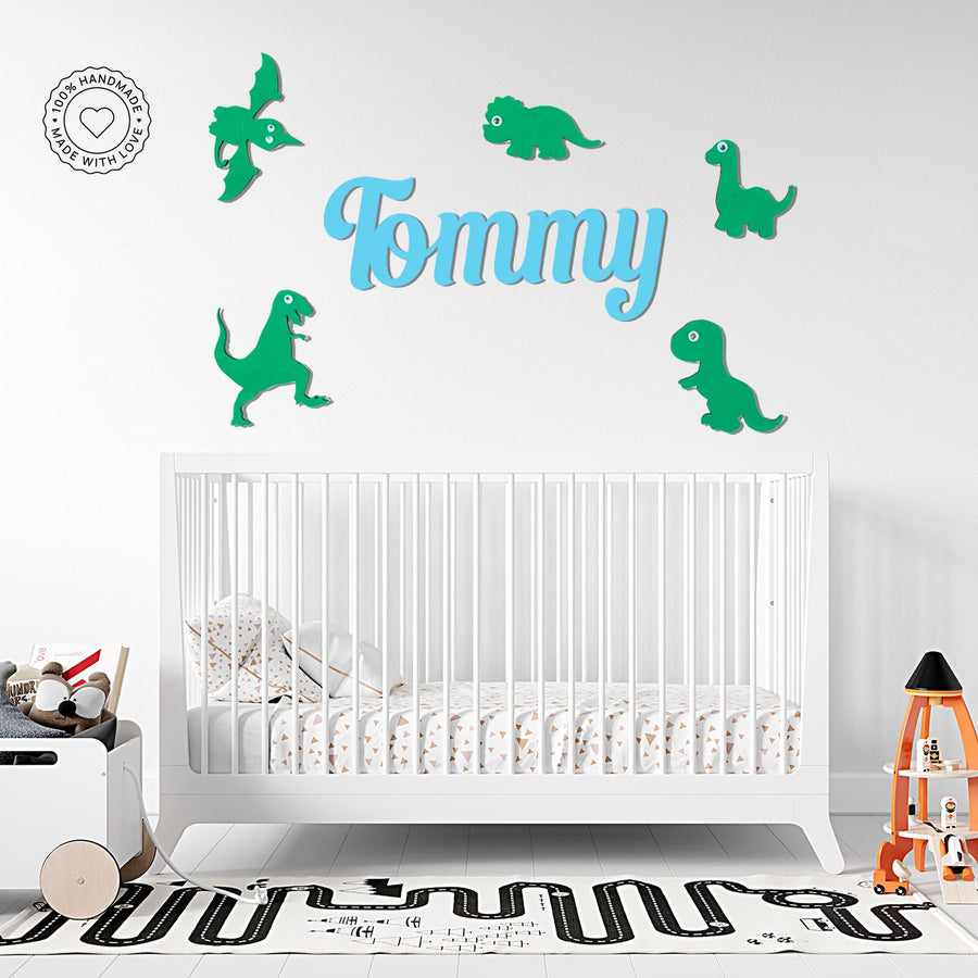 Personalised wooden name sign 'Tommy' in soft blue letters, surrounded by adorable green dinosaur characters on a white nursery wall. Five cartoon-style dinos including a T-Rex, long-neck, and pterodactyl frame the name, creating a playful prehistoric theme. The decorative wall art is mounted above a modern white crib with patterned bedding, adding a cheerful touch to this stylish nursery space.