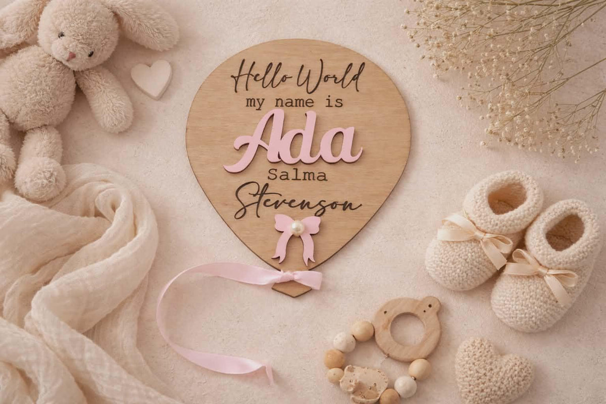 Hello World wooden balloon baby announcement plaque flat lay with pink ribbon and newborn accessories.