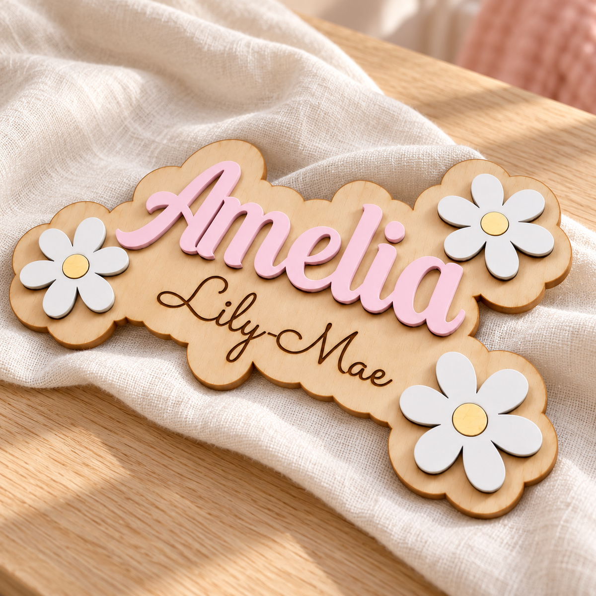 Angled view of the Amelia and Lily-Mae birch wood plaque, showing raised pastel pink lettering and layered white daisies.
