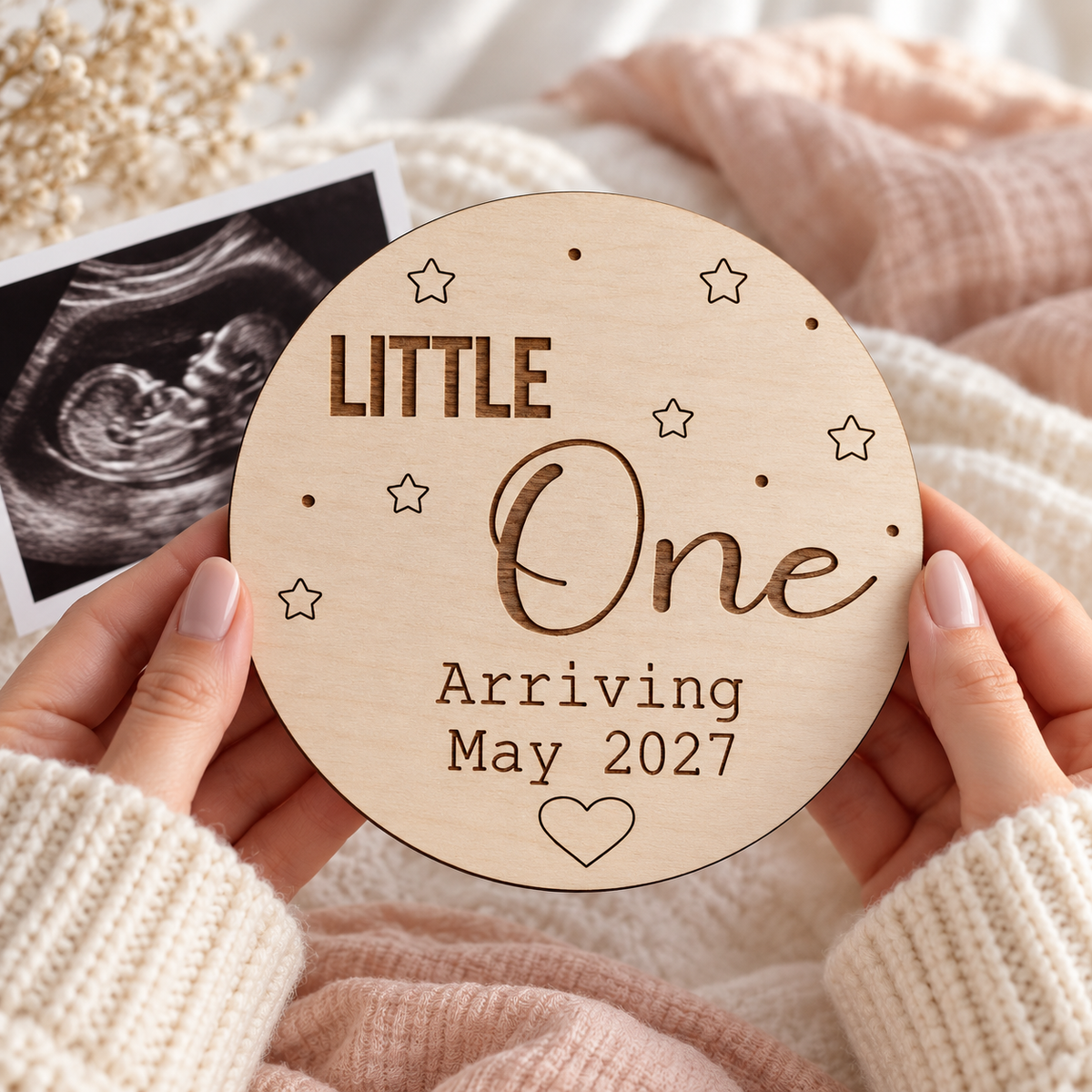 Hands holding a Little One Arriving engraved wooden pregnancy announcement sign beside an ultrasound