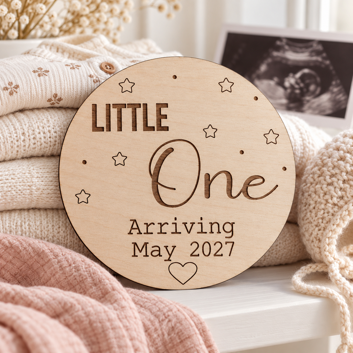 Engraved Little One Arriving plaque displayed beside folded cream baby clothes