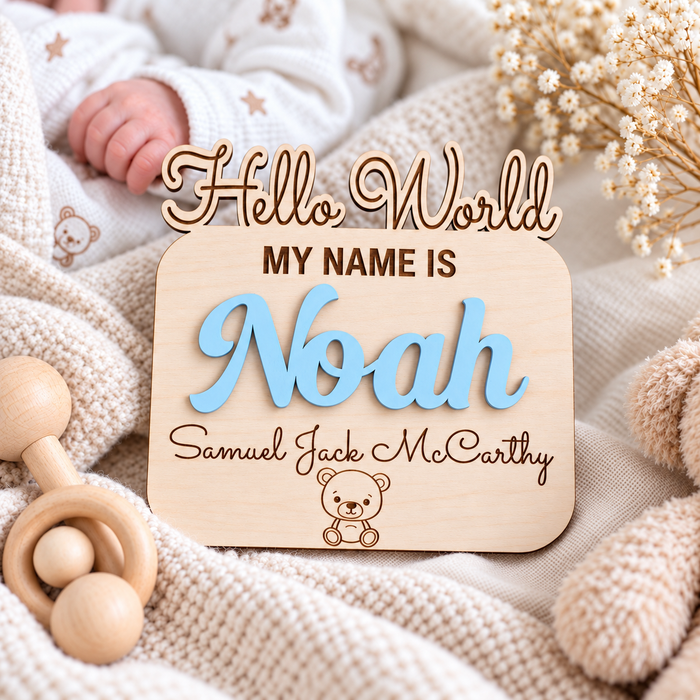 Personalised newborn name sign with Noah in raised Pastel Blue lettering, engraved extra names and a teddy detail beside a newborn's hand.