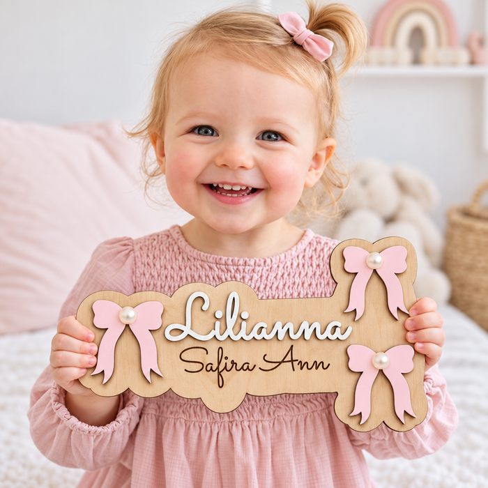 Personalised wooden bow name sign held by a smiling girl, with Lilianna in raised white lettering, Safira Ann engraved below and three pink pearl-centred bows.