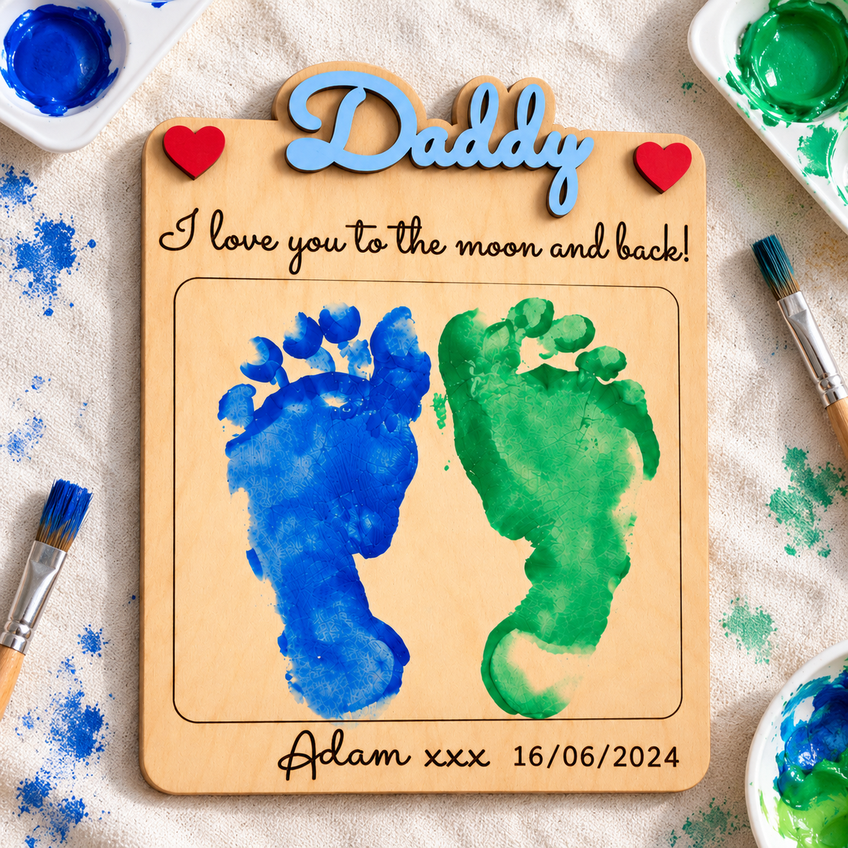 Personalised wooden footprint gift on a craft surface beside blue and green paints and brushes; paints are not included.