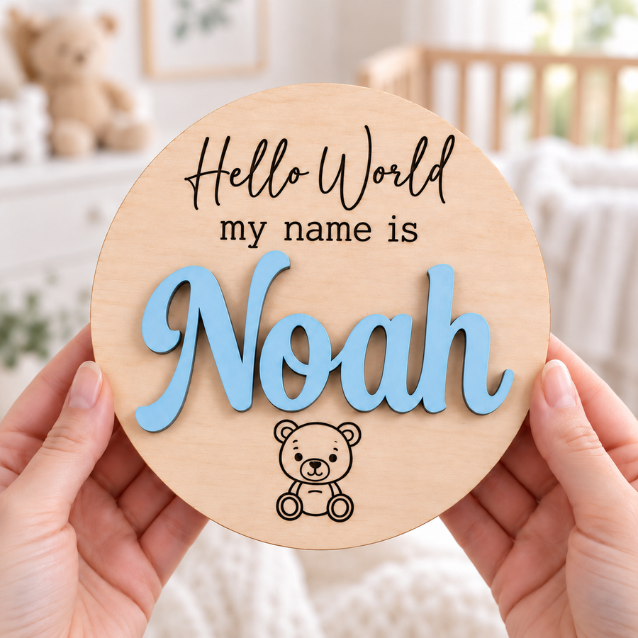 Personalised Hello World sign held in hands, with Noah in raised Pastel Blue lettering and dark engraved teddy bear on a round wooden plaque.