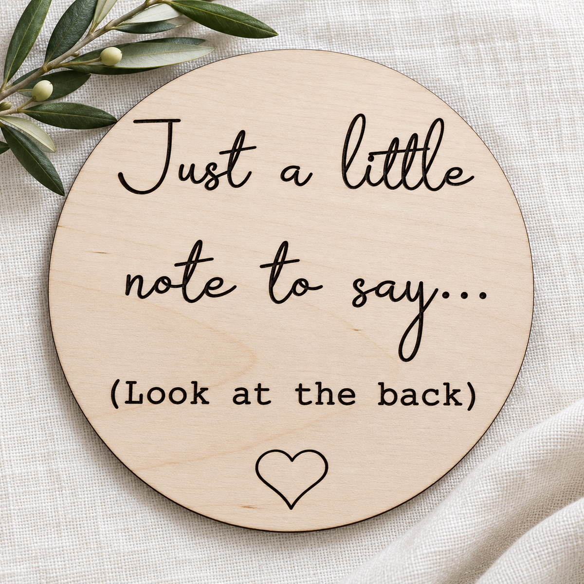 Front of the circular wooden pregnancy announcement plaque on linen beside an olive branch