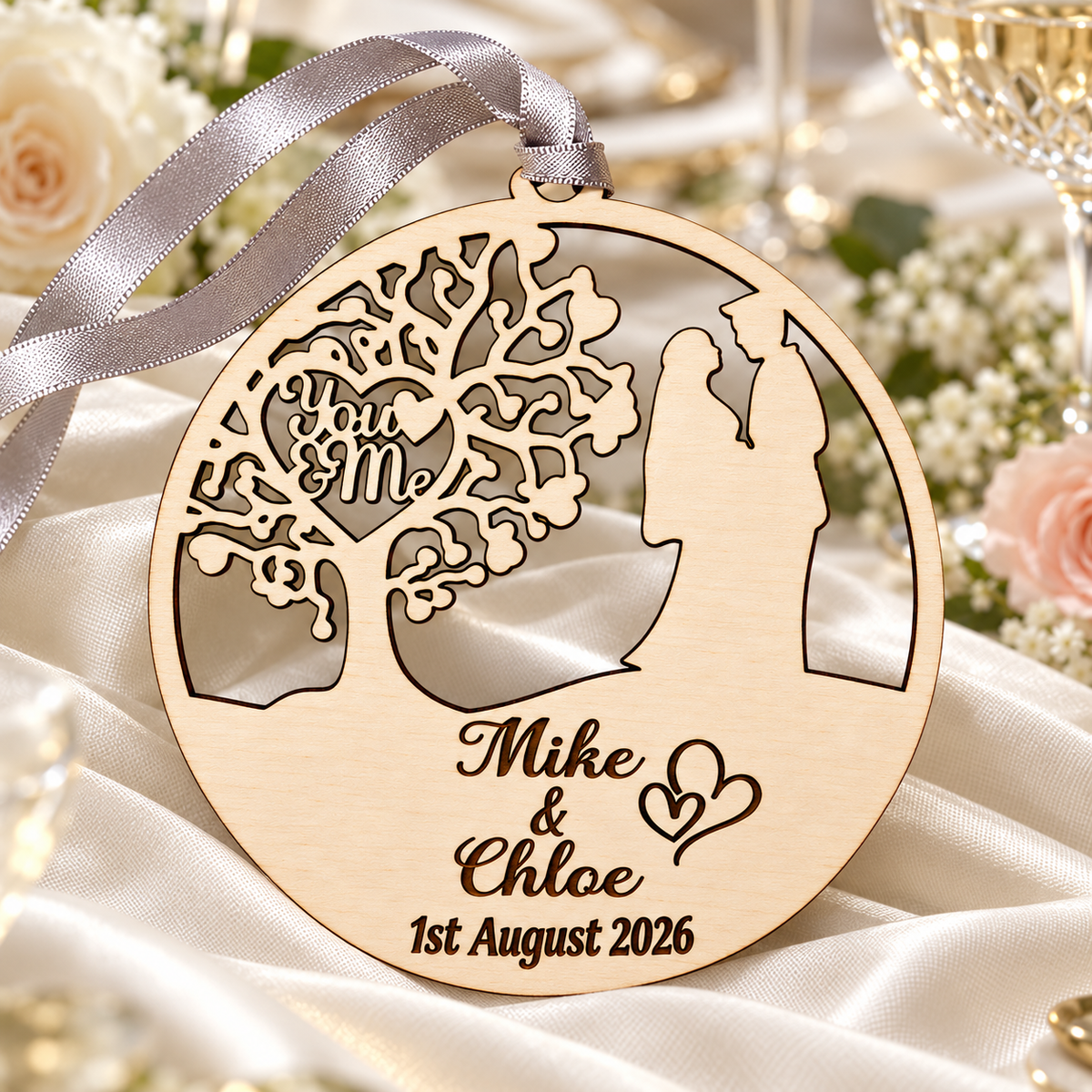Personalised You and Me wedding ornament styled on ivory fabric at a wedding table with flowers and glassware in the background.