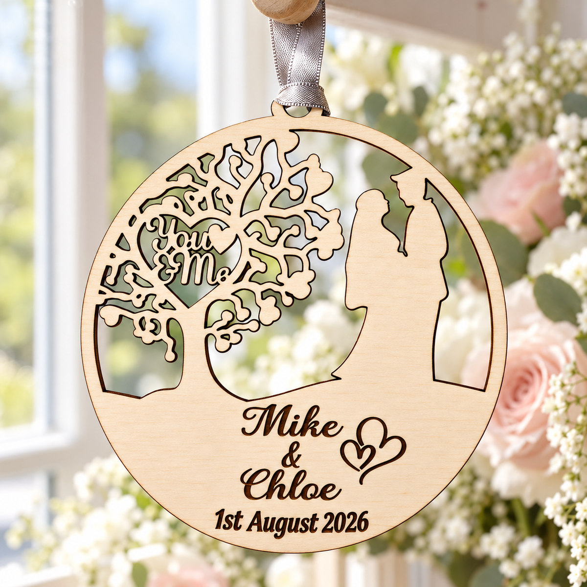 Personalised wooden wedding ornament hanging by a silver-grey ribbon beside a bright window, with You and Me tree cut-outs and dark engraved names and date.