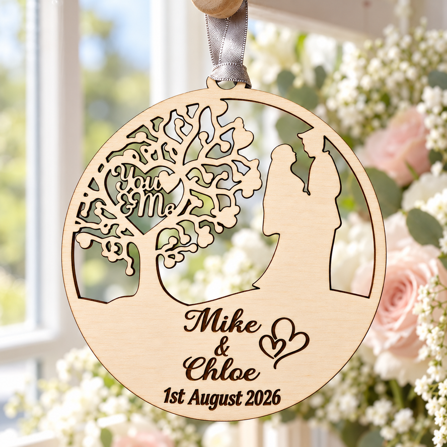 Personalised wooden wedding ornament hanging by a silver-grey ribbon beside a bright window, with You and Me tree cut-outs and dark engraved names and date.