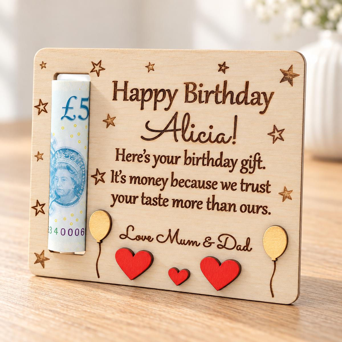 Close-up of a personalised wooden birthday money holder for Alicia with a rolled £5 note, engraved message, red hearts and yellow balloons