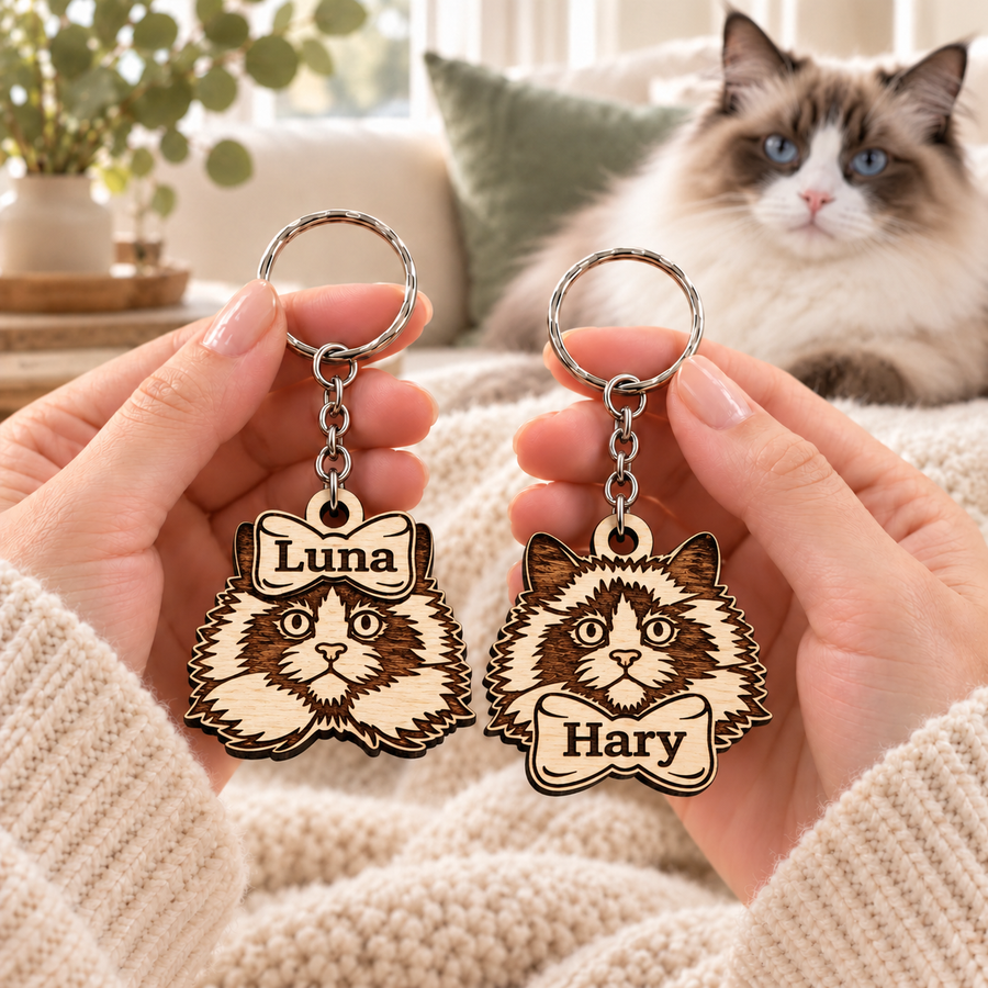 Personalised wooden Ragdoll cat keyrings with Luna on a top bow and Hary on a bottom bow, held beside a blue-eyed Ragdoll cat