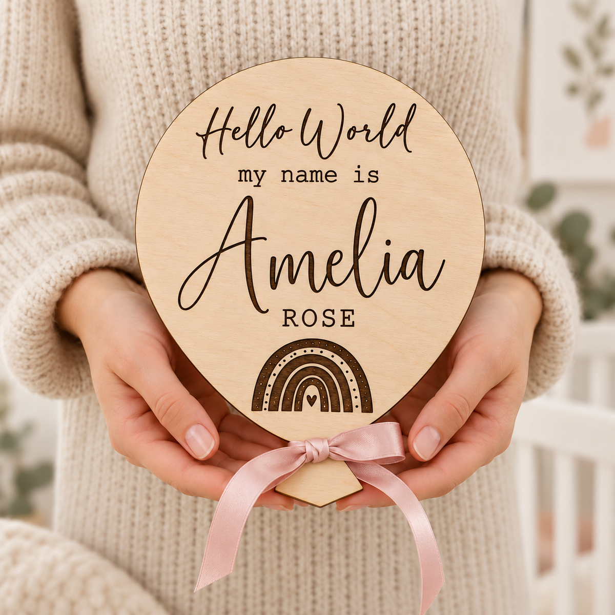 Engraved rainbow balloon baby name plaque held in adult hands showing the wooden shape and pink ribbon